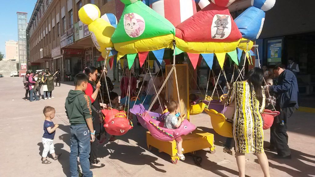 Images contrastées du Xinjiang : enfants qui font du manège avec, en fond, « les gardes de maison » du marché de Hotan, en train de pratiquer un exercice.