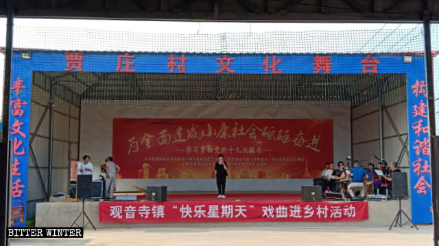 Un spectacle est en cours au centre culturel du village de Jiazhuang. Au-dessus de la scène, une banderole porte l’inscription « Dimanche heureux ».